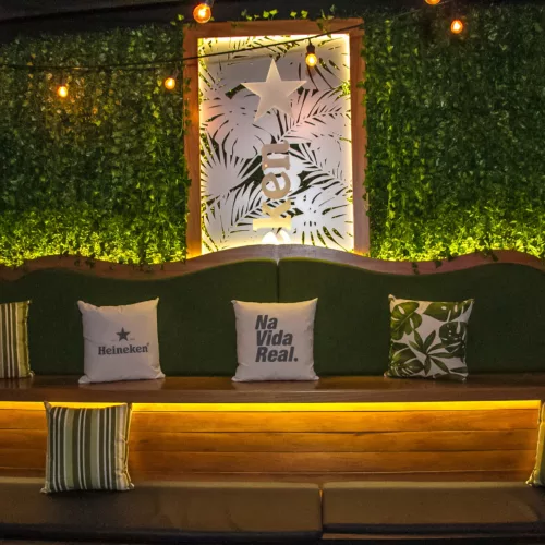 Espaço com parede verde iluminado e embaixo almofadas em um assento de madeira. Nas almofadas lê-se Heineken e o nome do local, Na Vida Real Bar.