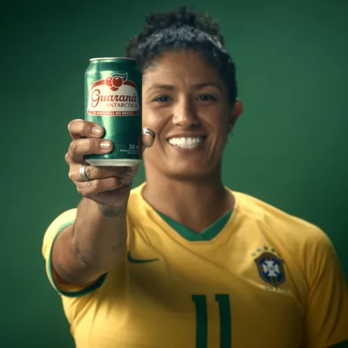 A jogadora Cristiane, com a camisa 11 da Seleção Brasileira Feminina de Futebol, segura uma lata de Guaraná Antarctica em frente a um fundo verde.