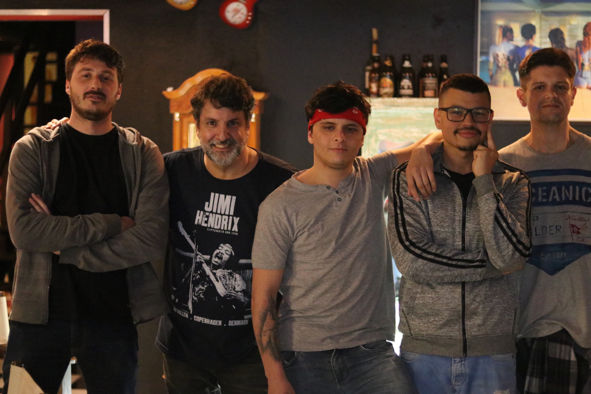 Tributo a Cazuza: cinco rapazes posam para a foto. O do meio usa um lenço caracterizando-se como Cazuza. Atrás vê-se um ambiente de bar.