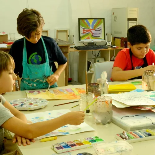 Crianças pintam e desenham com vários materiais sobre a mesa. Oficina Ateliê dos Arteiros.