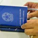 Pessoa manuseia carteiras de trabalho, na cor azul. Foto usada para ilustrar vagas de emprego em agências do Sine ou contratação por empresas.