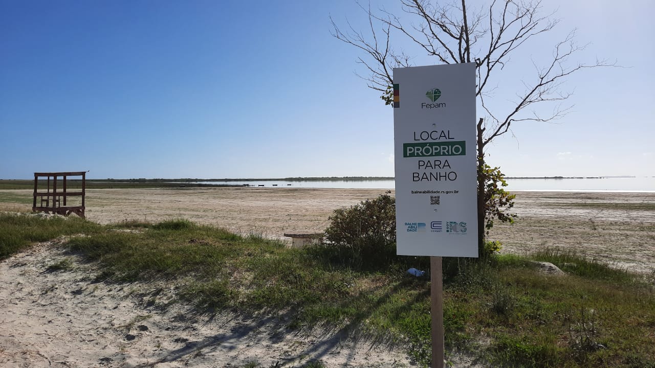 Placa da Fepam com a inscrição "local próprio para banho". Ao fundo, praia vazia e céu azul compõem a paisagem.