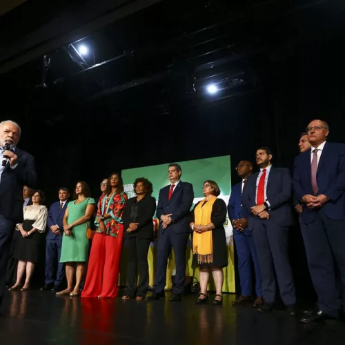O presidente eleito, Luiz Inácio Lula da Silva, em um palco, anuncia novos ministros que comporão o governo. Os novos ministros também estão no palco e observam o novo presidente da República falar.