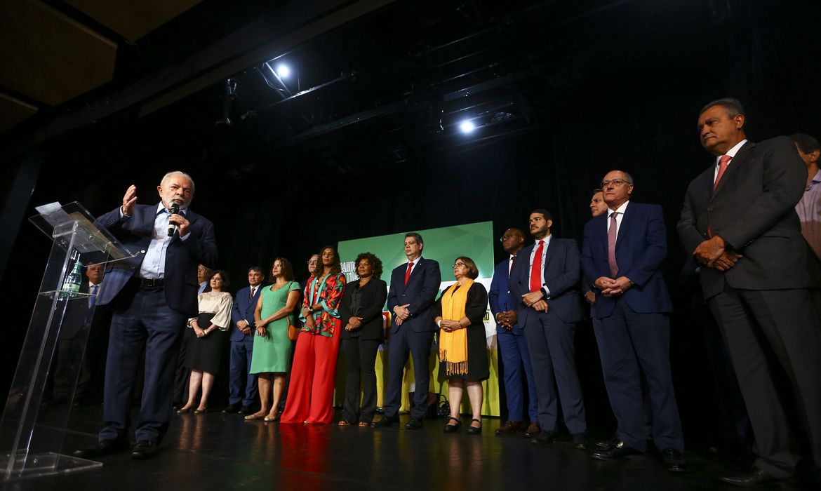 O presidente eleito, Luiz Inácio Lula da Silva, em um palco, anuncia novos ministros que comporão o governo. Os novos ministros também estão no palco e observam o novo presidente da República falar.