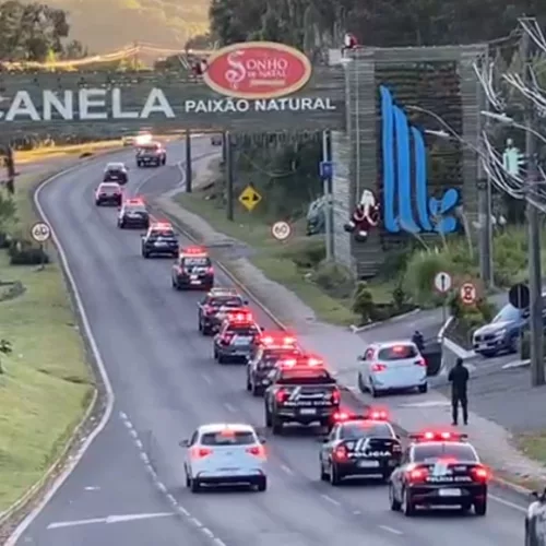 Treze viaturas da Polícia Civil estão em fila, andando em uma estrada. Elas estão passando pelo pórtico de entrada de Canela, na Serra do RS.