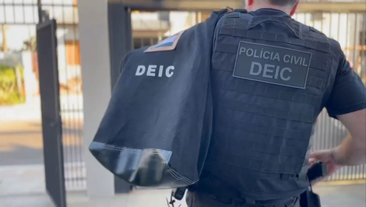 Policial civil veste um colete à prova de balas e carrega um malote com a expressão "DEIC". Ele está de costas e é possível ler "Polícia Civil DEIC". Ao fundo, é possível ver uma grade e um portão residencial.