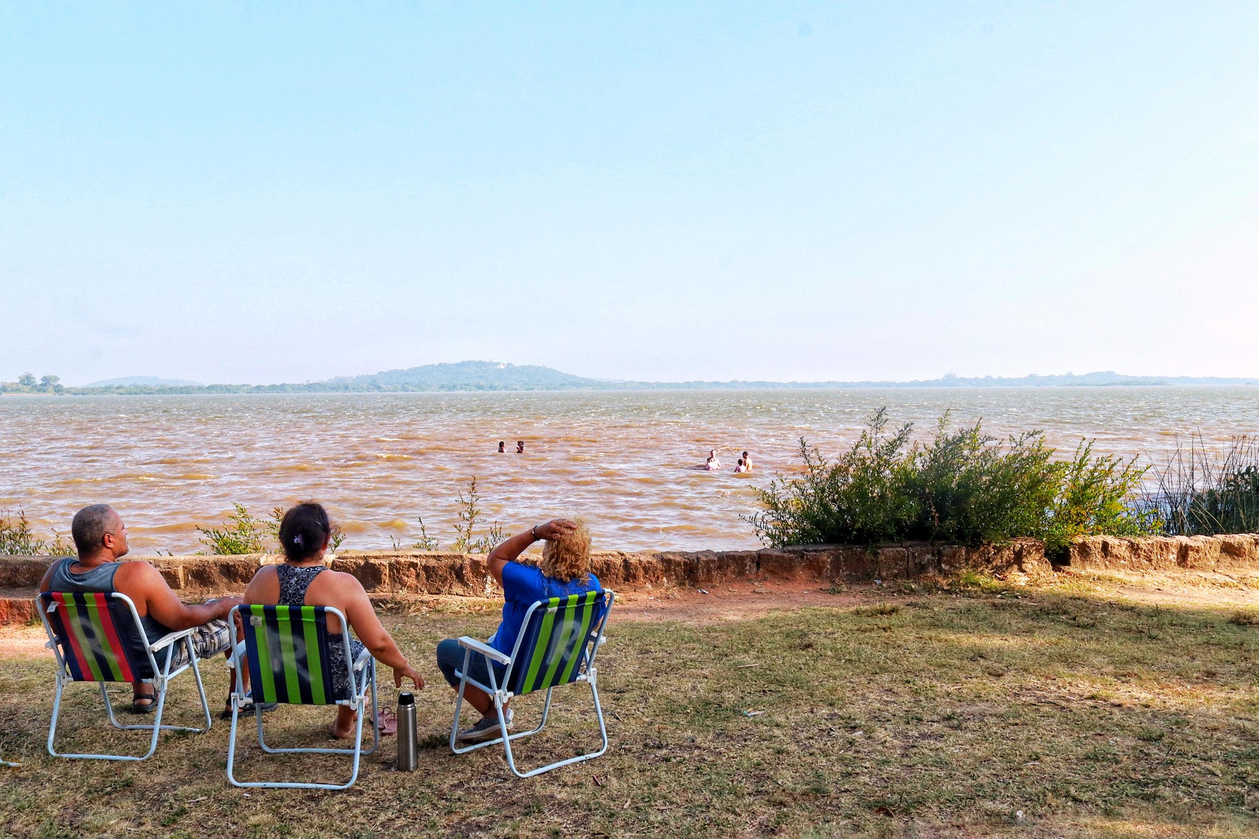 Três pessoas sentadas em cadeiras de praia contemplam a praia do Leblon, na Orla do Guaíba, em Porto Alegre. Ao fundo, o lago Guaíba.