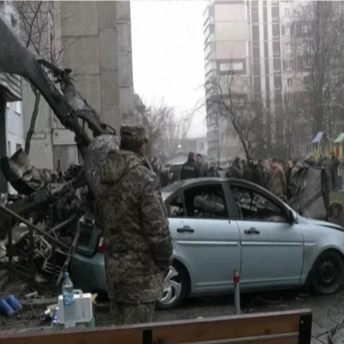 Soldado ucraniano olha para destruição causada em um prédio e um carro sedan prata após queda de helicóptero. Ao fundo, outras pessoas assistem a cena.