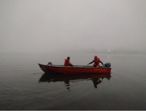 Barco tripulado por dois bombeiros de uniforme vermelho em meio às aguas do Rio Uruguai. O tempo está com neblina